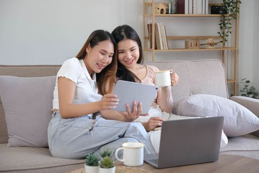 Two happy Asian women best friends in casual wear laughing while working with tablet at home in living room..