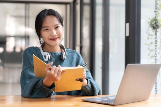 Young adult happy smiling Asian student wearing headphones talking on online chat meeting using laptop in university campus or at virtual office. College female student learning remotely