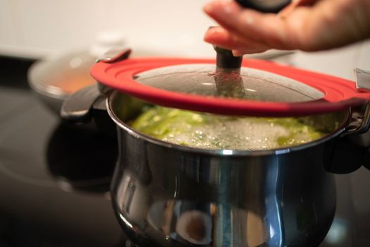 Close up of female hand opening pan lid on electric hob with boiling water with bubbles on green vegetable soup. Homemade tasty economic cooking concept.