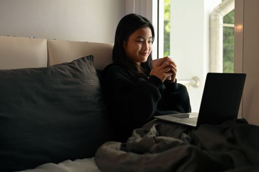 Smiling asian girl drinking hot chocolate and using laptop on bed, relaxing on weekend morning.