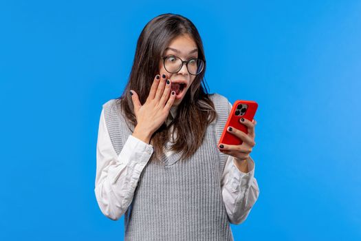 Surprised woman received surprise message on smartphone. Young woman with open mouth on blue background. . High quality photo