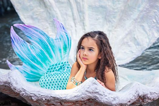A beautiful little brunette girl in a mermaid costume lie in a large sea shell, outdoor, looking away. Close up. Copy space