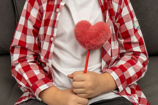 A cute boy is sitting on the couch in a white T-shirt and a plaid shirt and holding a fluffy toy in the shape of a heart. A manifestation of love. mothers Day. Father's Day. Thanksgiving day. valentine's day. close-up