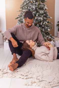 The girl, closing her eyes, lies on the lap of a stylish guy near the Christmas tree in the room.Vertical