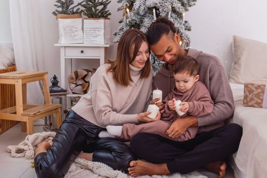 A young multi-racial family, in cozy sweaters, with a little boy sitting on a knitted blanket near the Christmas tree, in the white room, holding large candles, smiling