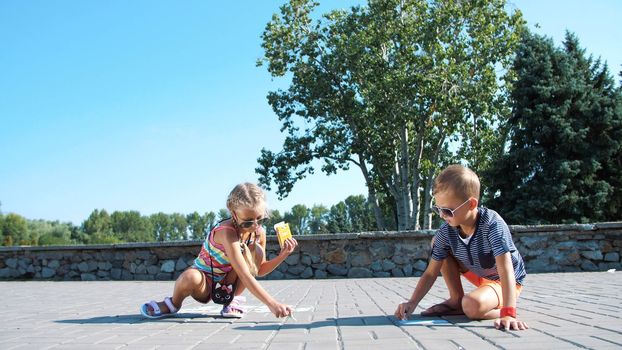 Children, a boy and a girl in sunglasses, paint with colored crayons on the asphalt, street tiles. A hot summer day. High quality photo