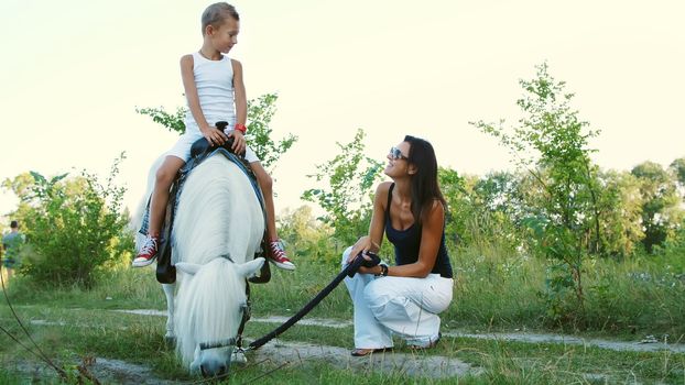 A woman and a boy are walking around the field, son is riding a pony, mother is holding a pony for a bridle. Cheerful, happy family vacation. Outdoors, in summer, near the forest. High quality photo