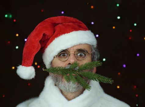 a man with glasses wearing a Santa Claus hat and a Christmas tree branch in his mouth on black with colored lights background. High quality photo