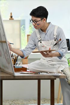 Portrait of asian male artist sitting in front of canvas and painting picture with watercolor.