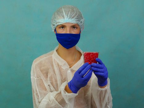 A guy in a robe, mask, gloves holds a glass of pills.Healthy lifestyle and young people, vitamins and food Supplements. A guy in blue gloves and hat and a white coat is holding a glass of red pills against a studio background.