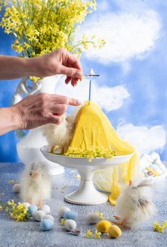 a woman lights a candle on a traditional yellow Slavic Easter curd cake on a blue background against blue sky with white clouds, High quality photo