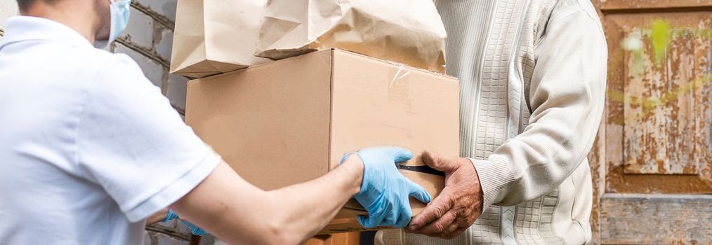 Young delivery man in uniform giving a box, parcel of groceries to elderly man outdoor. Shopping help and delivery service. Volunteer support seniors during coronavirus outbreak.