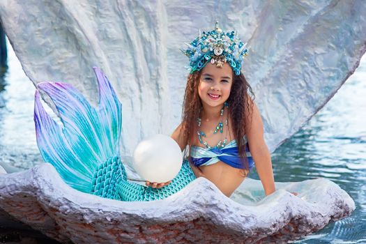 A beautiful happy little brunette girl in a mermaid costume and crown, sits in a large sea shell, outdoor, hold a big pearl . Close up. Copy space