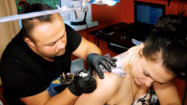 in a tattoo salon , a specialist is doing a tattoo on woman's back, a floral ornament. a man works in special gloves, on special equipment. drawing is done with black paint. High quality photo