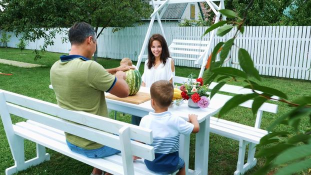 A young family with two little funny children sitting at a dinner table in the garden, in summer. Dad cuts a large watermelon, is going to treat his family. lunch with the family. High quality photo