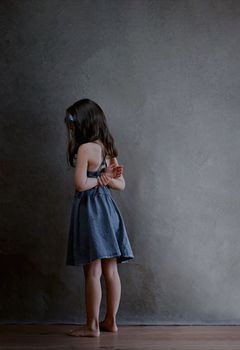 punished girl stands in the corner and is offended by the whole world, rear view of a girl with long dark hair in a blue summer dress holding her hands behind her back on a dark background. High quality photo