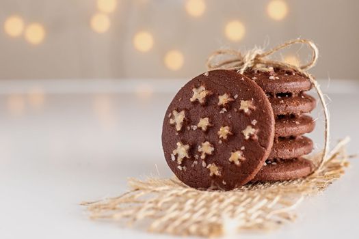 on a white table is a stack of round chocolate Christmas cookies with sugar stars and salt, tied with a rope, against a backdrop of shining garlands.