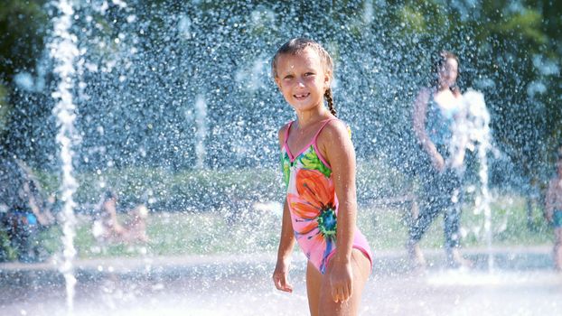 Happy girl of seven years, in swimsuit, having fun between water splashes in fountain . Sunny summer day in the city. Slow motion. High quality photo