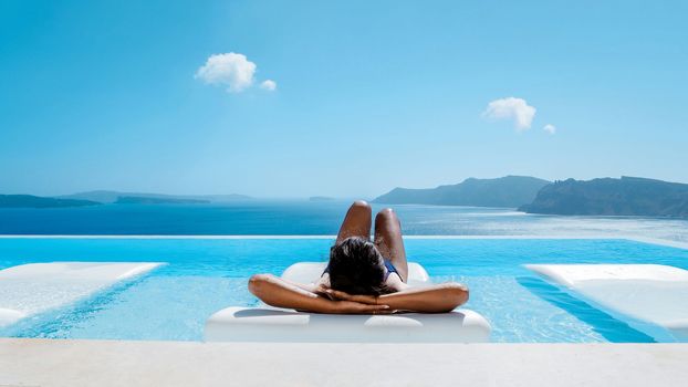 Young multicultural Asian woman in the Infinity swimming pool looking out over the ocean caldera of Oia Santorini Greece. women sun tanning in a pool with a blue sky