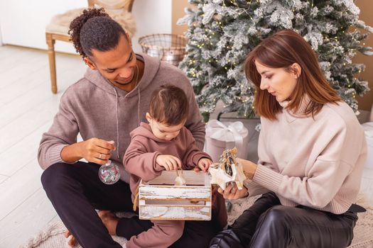A young stylish multi-racial family with a little boy near the Christmas tree, holding a wooden box with Christmas toys. Top view