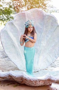 A beautiful little girl in a mermaid turquoise costume and crown is standing outdoors, in a large seashell, showing a heart with her hands. Vertical. Copy space