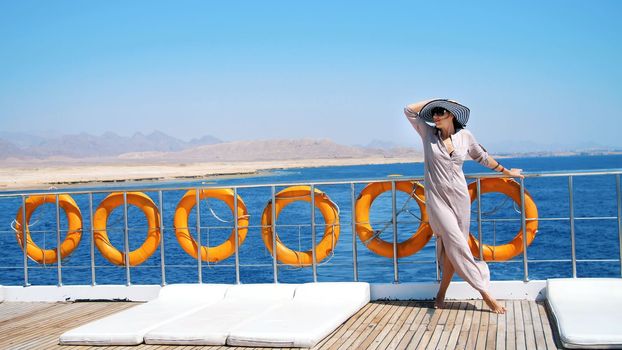 summer, sea, beautiful young brunette woman, in a long dress and sunglasses, stands on the deck of a ferry, ship, enjoying the rest, the beauty of the sea. on board hangs a lot of lifebuoys. High quality photo