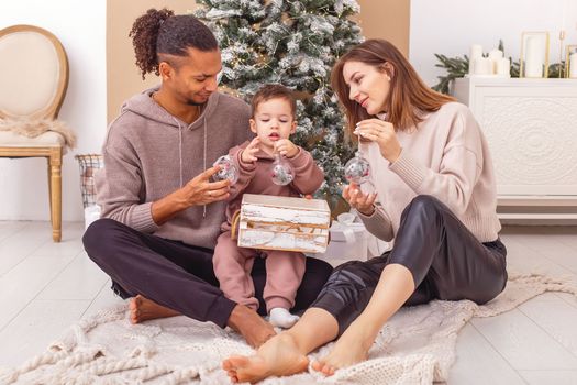 A happy multi-racial family sitting on a knitted blanket near the Christmas tree, holding a wooden box with Christmas toys.