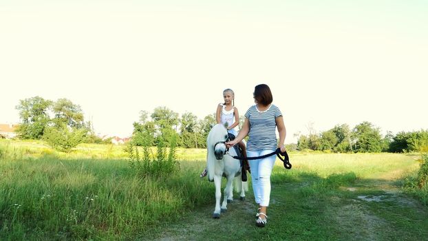 Mom and daughter are walking around the field, daughter is riding a pony, mother is holding a pony for a bridle. Cheerful, happy family vacation. Outdoors, in summer, near the forest. High quality photo