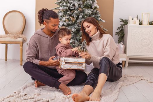 A happy multi-racial family with a little boy is sitting on a knitted blanket near the Christmas tree, holding a wooden box with Christmas toys.