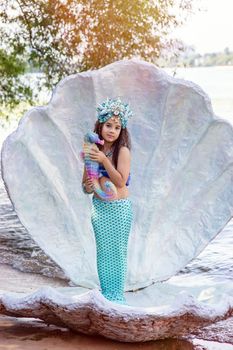 A smiling little girl in a turquoise mermaid costume and a crown is standing outdoors, in a large seashell, holding a colorful seahorse toy. Vertical. Close up. Copy space