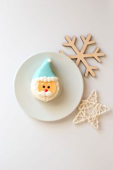 A cute sweet dessert in the form of a Santa Claus head, with a blue cap, lies on a white saucer, on a white table. Top view. Copy space. Vertical
