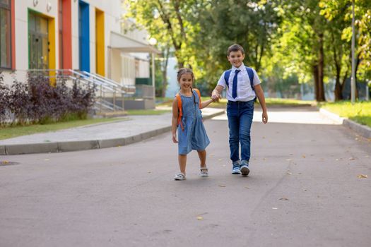 Cheerful schoolchildren, a girl and a boy, brother and sister with backpacks go from school in the morning on a sunny summer day against the background of a school building with multi-colored windows.