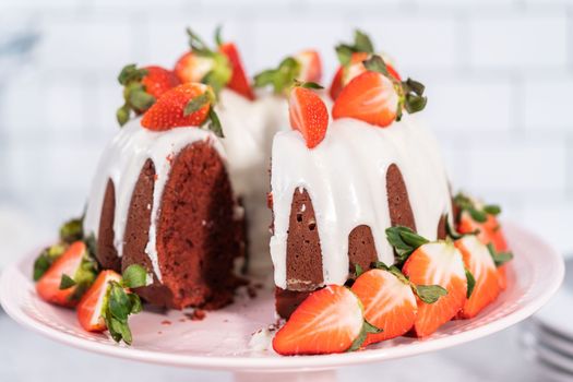 Slicing red velvet bundt cake with cream cheese frosting garnished with fresh strawberries.
