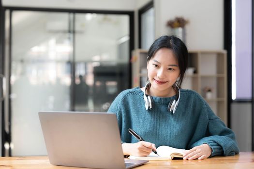 Young adult happy smiling Asian student wearing headphones talking on online chat meeting using laptop in university campus or at virtual office. College female student learning remotely