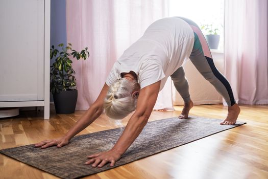 Senior woman working out at home, doing yoga exercise in the room downward facing dog pose, adho mukha svanasana from sun salutation pose. 