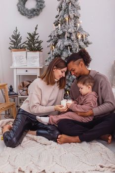 A young multi-racial family with a child is sitting on a knitted blanket near a snow-covered Christmas tree, in a room, holding large candles in their hands. Vertical