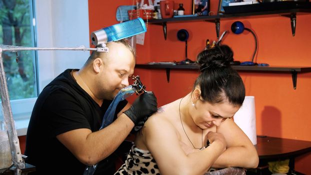 in a tattoo salon , a specialist is doing a tattoo on woman's back, a floral ornament. a man works in special gloves, on special equipment. drawing is done with black paint. High quality photo