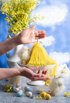 a woman lights a candle on a traditional yellow Slavic Easter curd cake on a blue background against blue sky with white clouds, High quality photo