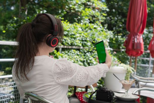 brunette girl listens to music in headphones with a green screen phone, at a table in a cafe, High quality photo