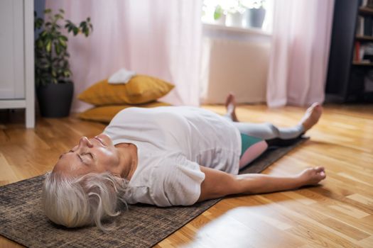 Senior caucasian woman lying on yoga mat after workout. Fit female relaxing on floor at home. 