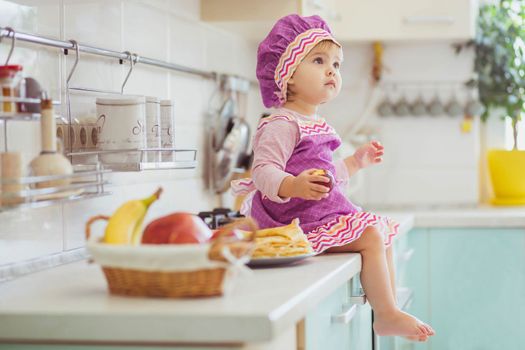 Adorable baby in an apron and chef's cap sitting with an apple on the table.