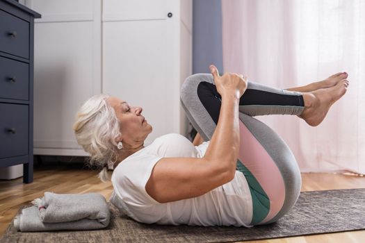 Senior woman lying on yoga mat doing apanasana. Fit female relaxing on floor at home. 