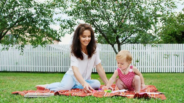 a young beautiful mother playing with a one-year-old daughter, in the garden, sitting on a plaid, a blanket, a coverlet, on the grass, lawn, in the summer. High quality photo