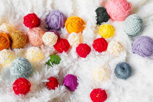Bright multicolored knitting yarn and a luminous garland in the form of balls lie on a white fluffy blanket. Cozy still life made of yarn. Close up.