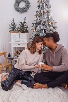 A young woman and a dark-skinned guy are sitting on a knitted blanket near a snow-covered Christmas tree in the room, holding large candles in their hands. Vertical