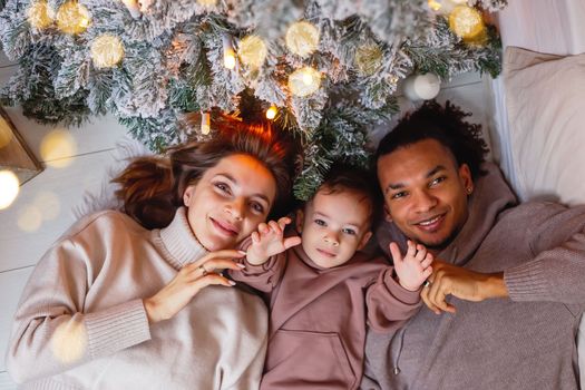 A happy young multiracial family with a little boy are lying on a knitted blanket, under a Christmas tree. Top view. Close up