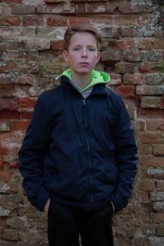 A 14 year old boy standing in front of an old brick wall. Boy standing against brick wall. Portrait of handsome teenager against old wall. Teenager posing and holding his hands in his pants pockets.