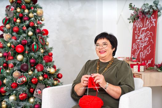 A cheerful 60 year old woman knitting a sock near the Christmas tree. elderly woman resting, relaxing during christmas holidays