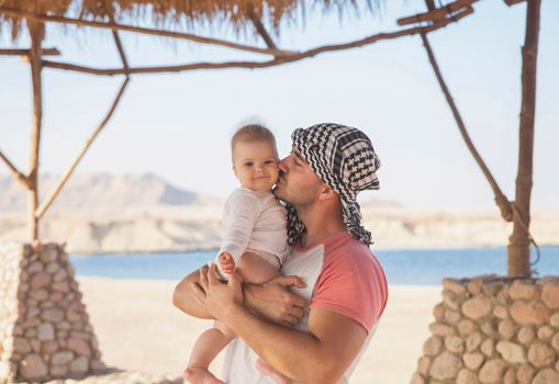 Father kisses his newborn daughter under a straw awning in the desert.
