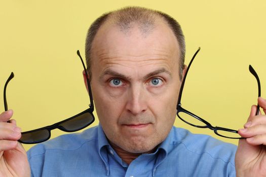 A handsome and intelligent man, 45-55 years old, with a yellow background, wearing a blue shirt, is holding two glasses and looking into the camera.